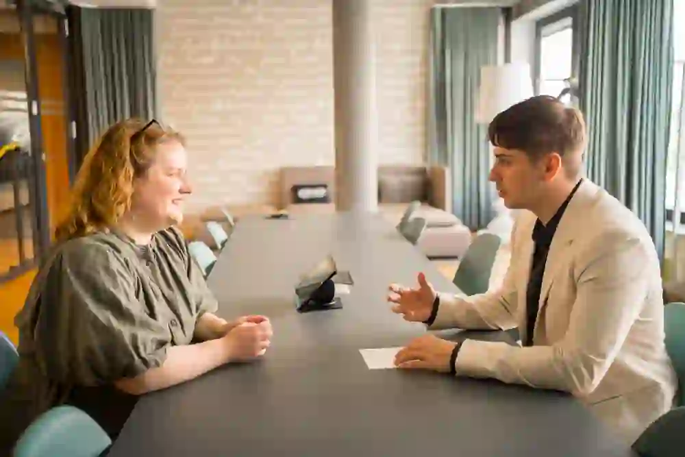 L'immagine mostra due persone sedute di fronte a un lungo tavolo che conversano. L'atmosfera è concentrata e amichevole. Entrambe le persone sembrano interessate e impegnate, con la donna che sorride e l'uomo che gesticola per sottolineare i suoi punti. L'atmosfera è professionale e piacevole, supportata da un arredamento luminoso e moderno.