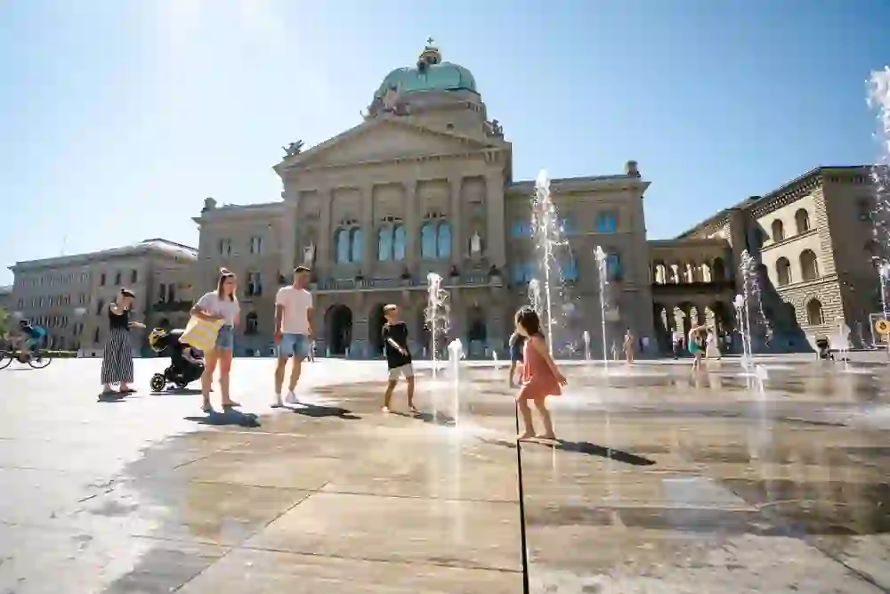 Devant le Palais fédéral à Berne, des personnes profitent de la journée ensoleillée tandis que des enfants jouent joyeusement entre les fontaines bouillonnantes - une image du bonheur estival.