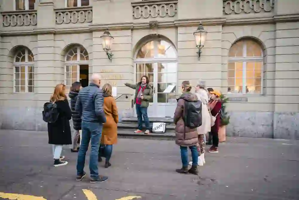 Ein Stadtführer in Bern steht vor einem historischen Gebäude, gestikuliert und erzählt Geschichten, umgeben von einer Gruppe interessierter Zuhörer, eingehüllt in ihre Jacken gegen die Kühle, gefesselt von den Ausführungen zur Geschichte ihrer Stadt auf der „1848 – Bern wird Bundesstadt“ Tour.