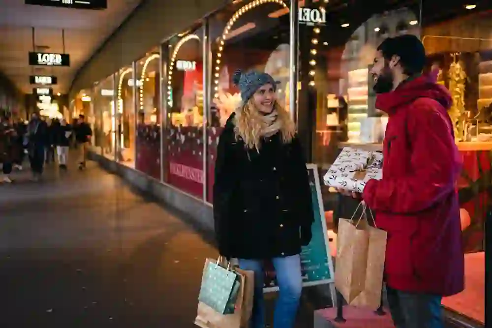 La photo montre deux personnes faisant du shopping de Noël devant une vitrine décorée pour la fête. Elles rient et tiennent des sacs cadeaux dans leurs mains. L'ambiance est joyeuse et festive, avec un soupçon d'anticipation des fêtes de fin d'année.