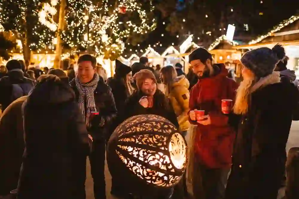 Das Bild zeigt eine Gruppe von Menschen auf einem Weihnachtsmarkt, die fröhlich zusammenstehen und warme Getränke genießen. Umgeben von festlicher Beleuchtung und dekorierten Ständen herrscht eine gemütliche und feierliche Stimmung.