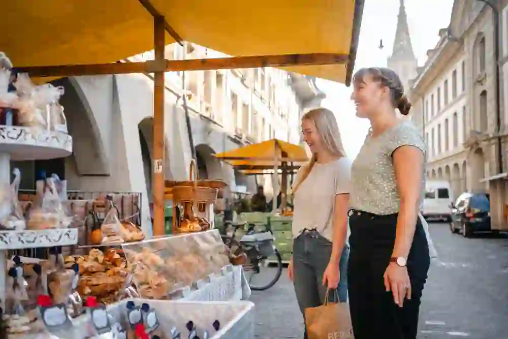 La photo montre deux jeunes femmes qui se tiennent devant un stand de pâtisseries sur un marché hebdomadaire. Elles sourient et semblent de bonne humeur en regardant les différents pains, pâtisseries et autres gourmandises. L'étal du marché a un store jaune et les marchandises sont présentées de manière attrayante, parfois emballées dans des sacs ou exposées dans des paniers.  En arrière-plan, on peut voir des bâtiments historiques avec des arcades, typiques de la vieille ville de Berne. On distingue également un clocher, probablement celui de la cathédrale de Berne. Les rues sont pavées et d'autres stands de marché sont visibles à l'arrière-plan. La scène semble conviviale et accueillante, avec une chaude lumière du soleil qui éclaire les environs.