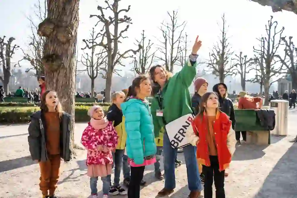 Un gruppo di bambini ascolta affascinato una guida della città in un parco di Berna. Gli occhi luminosi e gli sguardi attenti testimoniano la meraviglia e la curiosità dei bambini, che stanno imparando giocando alla luce del sole.