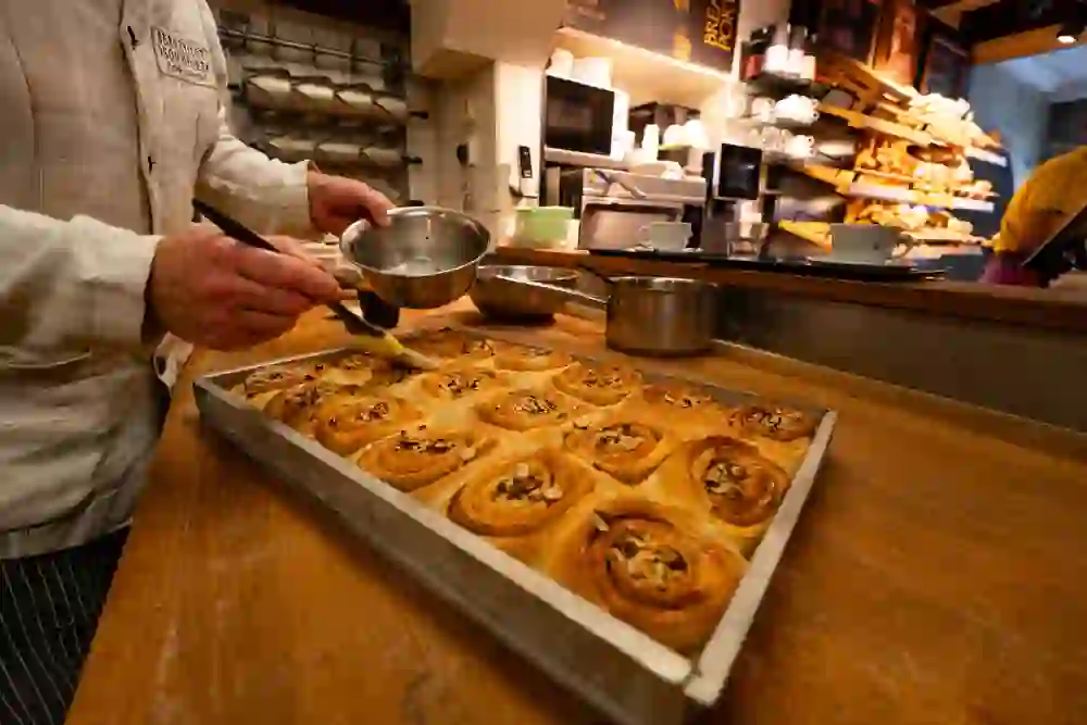In der Backstube von Bread à Porter werden frisch gebackene Zimtschnecken liebevoll glasiert. Der goldene Teig glänzt im warmen Licht, während der Duft von Zimt und Nüssen eine gemütliche, handwerkliche Atmosphäre voller Genuss und Hingabe verströmt.