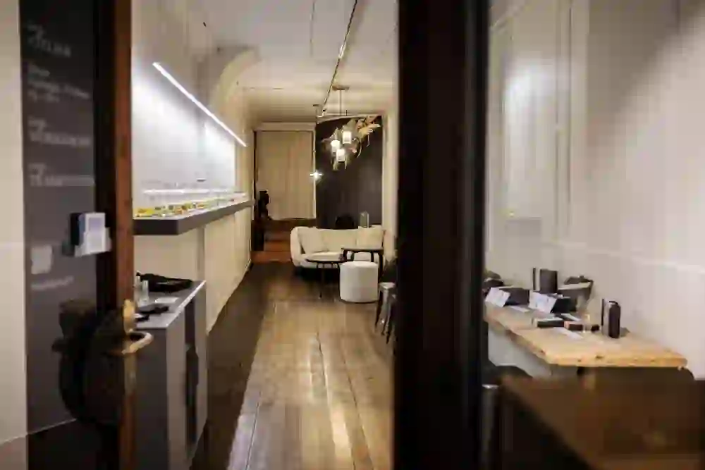 Das Bild zeigt einen stilvoll eingerichteten Raum, der vermutlich für Workshops oder Duftverkostungen genutzt wird. Der lange Holzboden und die warmen Farbtöne der Einrichtung schaffen eine einladende und gemütliche Atmosphäre. An der linken Wand befindet sich ein Regal mit Fläschchen, wahrscheinlich Duftöle oder Essenzen, die ordentlich aufgereiht sind und zum Experimentieren einladen. Im Hintergrund stehen bequeme Sessel und ein kleiner Tisch, was auf einen Lounge-Bereich hinweist. Die Raumgestaltung wirkt durchdacht und lädt die Besucher ein, sich zu entspannen und das sensorische Erlebnis in einer ruhigen Umgebung zu genießen.