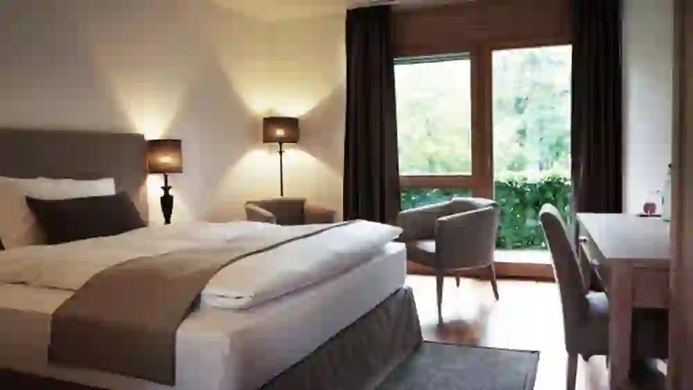 La photo montre une chambre d'hôtel moderne et confortable au château de Hünigen, avec une grande fenêtre qui donne sur la verdure. L'éclairage chaleureux et l'aménagement accueillant donnent un sentiment de calme et de confort.