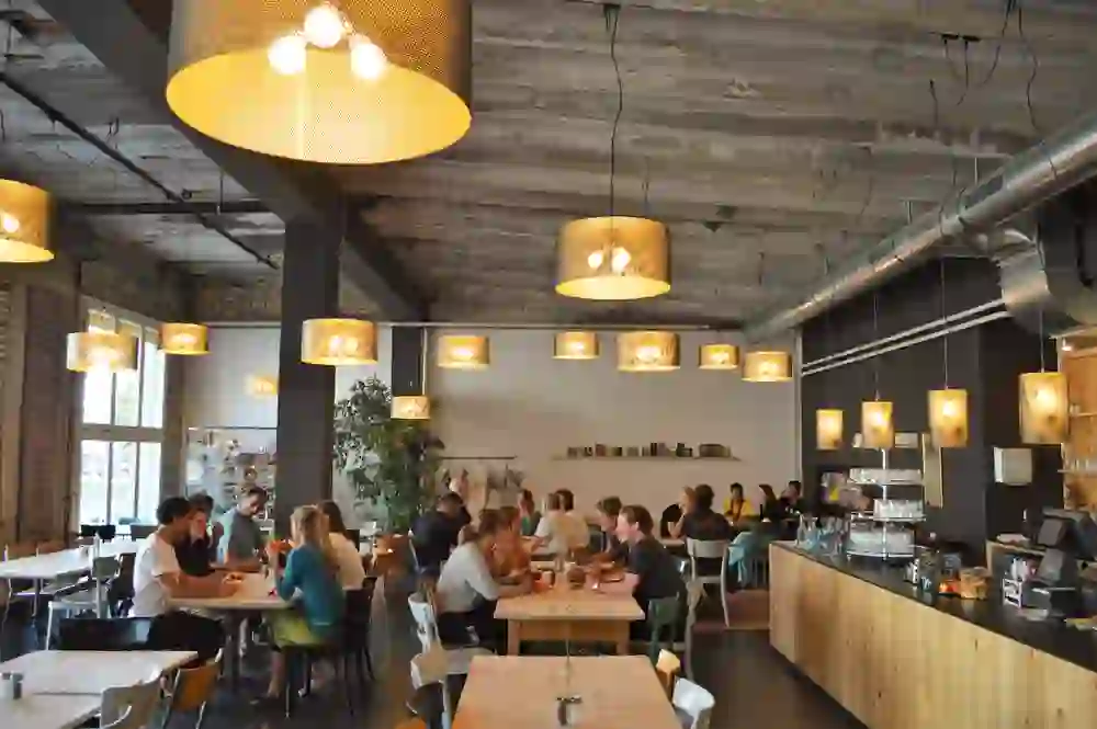 Das Bild zeigt ein belebtes Café mit einer rustikalen, modernen Atmosphäre. Menschen sitzen an Tischen und genießen Gespräche und Essen, umgeben von warmem Licht und stilvollen Hängelampen. Die offene, freundliche Gestaltung lädt zum Verweilen und Genießen ein.