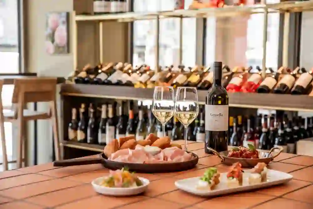The picture shows a table laid with a selection of Italian antipasti, accompanied by a glass of white wine and a bottle of wine. Wine bottles can be seen in the background, suggesting a wine bar. The atmosphere is inviting and cosy, perfect for an enjoyable meal in relaxed surroundings.