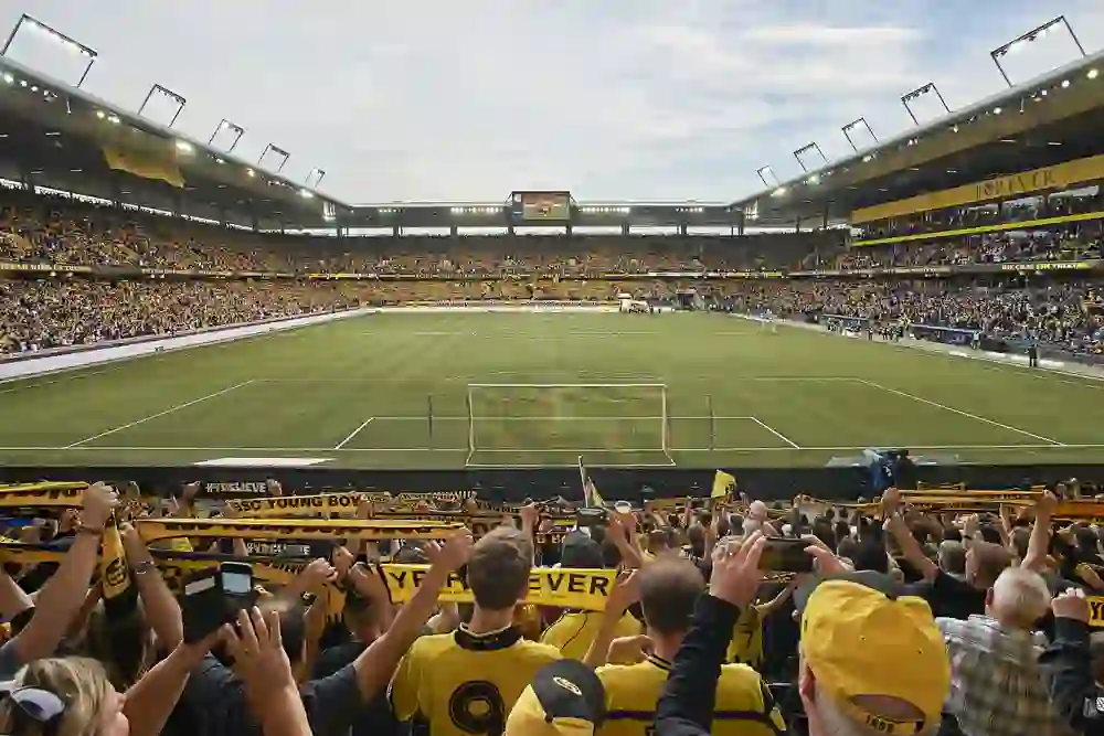 Das Bild zeigt das Wankdorfstadion während eines Fußballspiels, mit einer lebendigen Atmosphäre und gefüllt mit Fans, die in gelb und schwarz gekleidet sind, die Farben des BSC Young Boys. Es scheint ein Heimspiel zu sein, da die Fans Banner mit dem Namen des Clubs halten. Die Zuschauertribünen sind gut besetzt, und die Menschenmenge sieht engagiert und enthusiastisch aus, viele mit erhobenen Armen, was auf eine positive Stimmung im Stadion hindeutet. Im Hintergrund ist ein Teil des Spielfeldes zu sehen, das von der Menge umgeben ist, und über den Tribünen ist eine elektronische Anzeigetafel sichtbar. Es ist ein Tagesspiel, da das Stadion hell und natürlich beleuchtet ist.