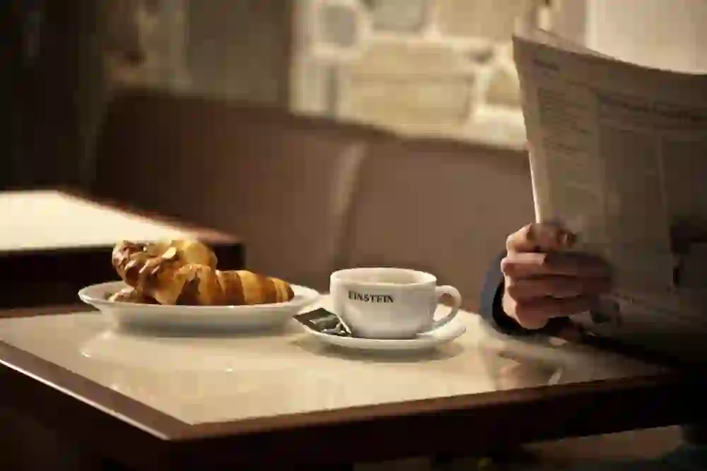 Auf dem Bild ist ein mit Kaffee und Croissant gedeckter Tisch im Einstein café & bel étage zu sehen. Am Tisch sitzt eine Person mit einer Zeitung.