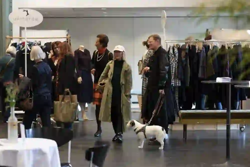 L'immagine mostra una sfilata di moda o un mercato della moda in un interno. Diverse persone guardano i vestiti esposti sugli stendini. In primo piano, una donna indossa un cappello chiaro e un cappotto verde. Accanto a lei c'è un uomo in giacca nera con un cane bianco al guinzaglio. Sullo sfondo si vedono altre persone, manichini e scaffali di vestiti. Ci sono due cartelli con la scritta "aéthérée" e un altro nome non ben leggibile. L'atmosfera è vivace e interessata.