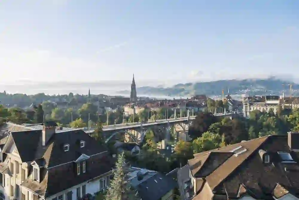 L'immagine mostra una vista pittoresca della città di Berna, con la suggestiva Cattedrale di Berna al centro e un ponte in primo piano. Sullo sfondo si ergono le colline, parzialmente avvolte dalla foschia mattutina. La foto emana uno stato d'animo limpido e tranquillo, con la luce del sole che illumina la città e crea un'atmosfera pacifica, quasi idilliaca. I tetti degli edifici in primo piano aggiungono un tocco tradizionale che ben si adatta all'atmosfera storica della città.