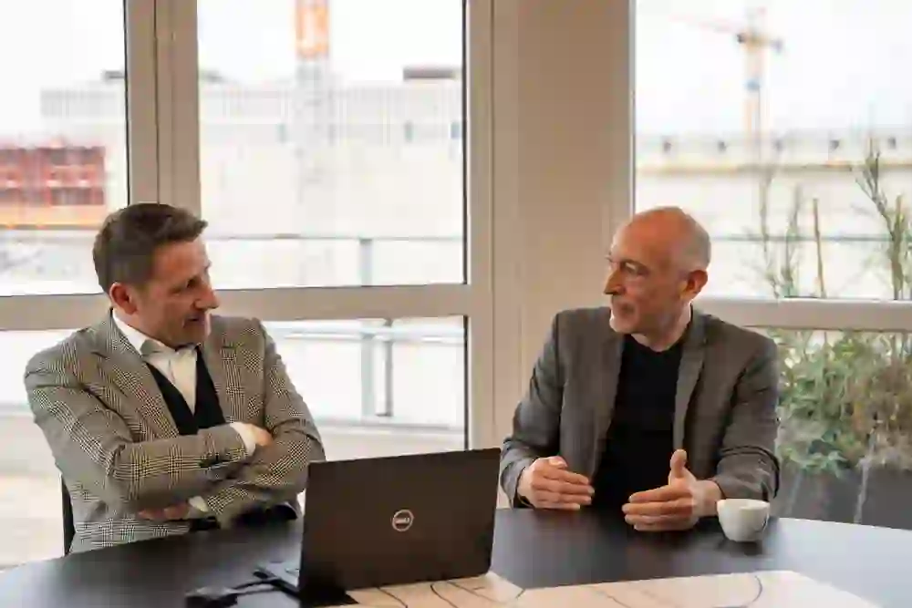 Deux hommes en tenue d'affaires sont assis à une table et discutent ; l'un sourit pendant que l'autre explique, dans une atmosphère qui suggère la collaboration et l'échange professionnels.