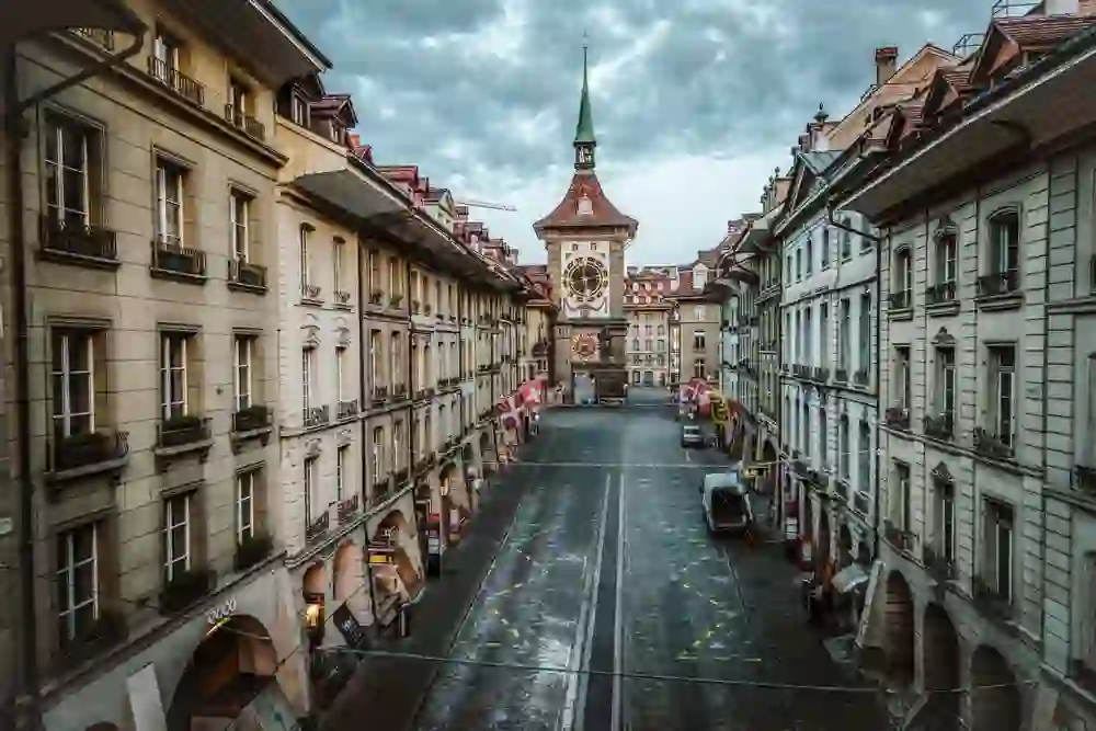 Auf dem Foto sieht man eine Straße in Bern, die zum Zytglogge, dem berühmten Uhrturm, führt. Die Straße ist gesäumt von traditionellen Gebäuden mit Balkonen und Fahnen, darunter die Schweizer Flagge.