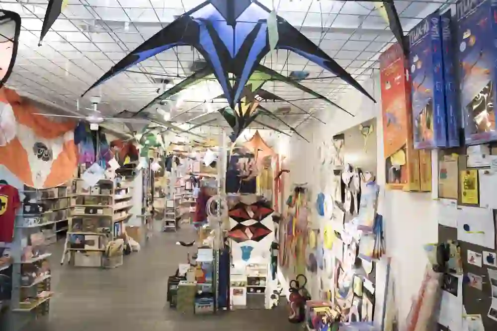 The picture shows the lively interior of a toy shop or a shop for leisure activities. Large, colourful kites are suspended from the ceiling, creating a playful atmosphere. The shelves are full of a wide variety of products, including games, books and leisure items that invite you to discover and browse.