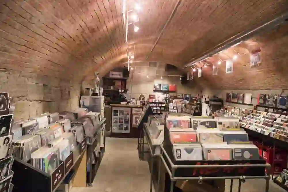 The photo shows the record shop Serge and Peppers Records, which is housed in a vaulted cellar. The cosy lighting and brick ceiling create a warm, inviting atmosphere. Carefully sorted records and music albums can be seen everywhere, suggesting a passionate dedication to music. One person is working at the counter in the background, giving the room a sense of busyness.