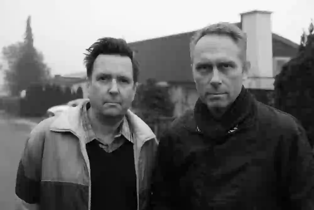 Two men look seriously into the camera - the mood is cool and thoughtful, as if a quiet moment were telling a larger story.