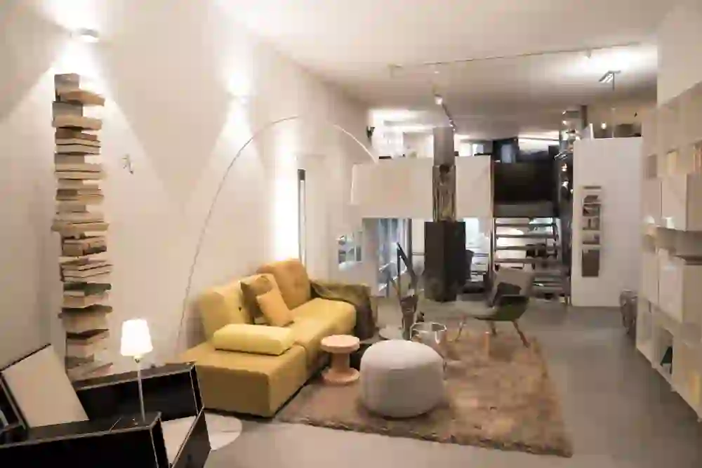 Das Foto zeigt den modernen, stilvoll eingerichteten Möbelladen Intraform. Ein gelbes Sofa bildet den Mittelpunkt, umgeben von einer harmonischen Kombination aus einer grossen Stehlampe, einem runden Couchtisch und einem bequemen Sessel. Eine Wand aus Büchern fügt eine kreative und intellektuelle Note hinzu. Die Beleuchtung ist warm und einladend, was die gemütliche Atmosphäre unterstreicht.