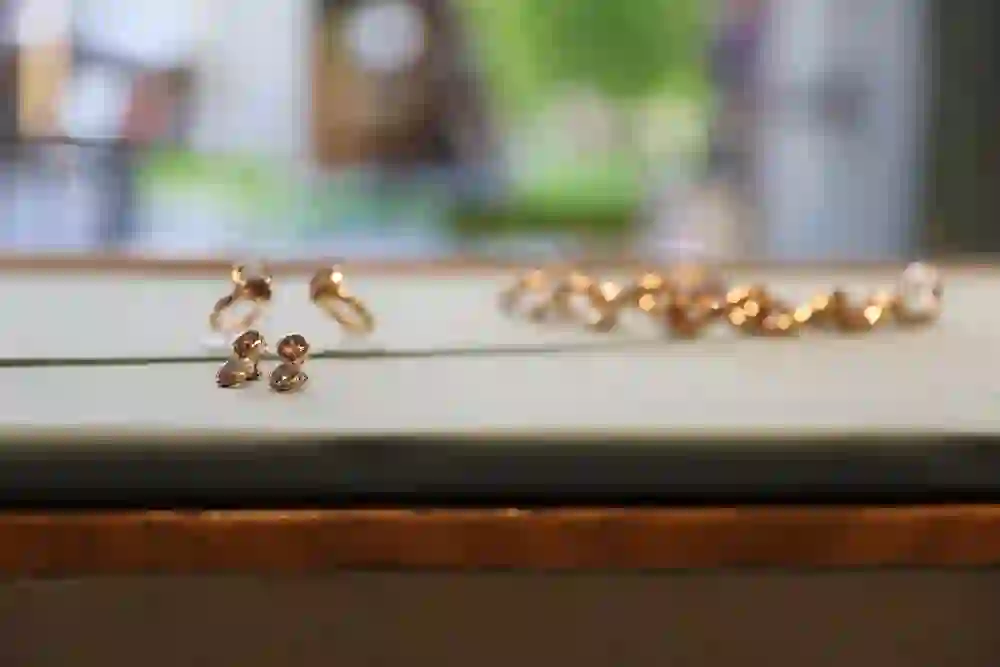 The picture shows elegantly presented pieces of jewellery on display in a jewellery shop. The focus is on earrings and rings, which are presumably made of gold and set with pink-coloured gemstones. A necklace can be seen in the background. The jewellery is presented on a surface with a discreet background, allowing the precious pieces to take centre stage.