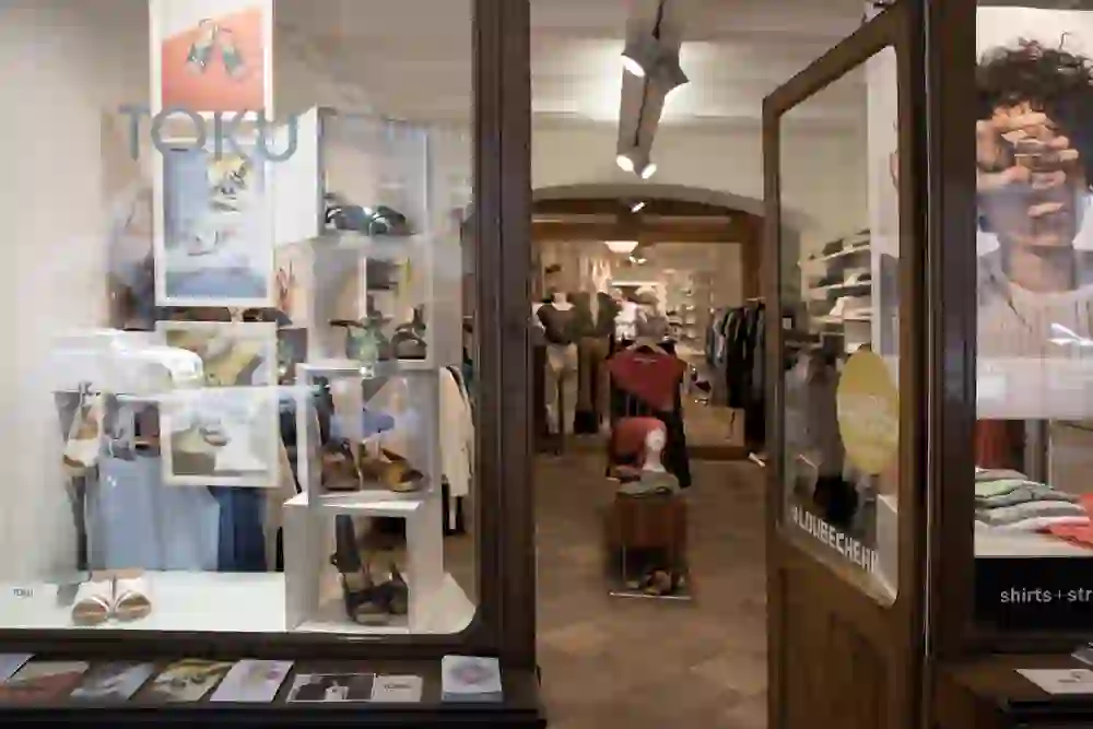 Das Bild zeigt den Blick durch die Eingangstür eines Geschäfts, wahrscheinlich eines Bekleidungs- oder Schuhgeschäfts, mit dem Namen "TOKU" deutlich sichtbar auf dem Schaufenster. Im Schaufenster sind verschiedene Schuhe ausgestellt. Im Inneren des Ladens sind Kunden und Kleidungsstücke zu sehen, die an Kleiderständern aufgehängt sind. Der Laden hat ein modernes und einladendes Ambiente mit einer Holzdecke und passenden Bodenbelägen.