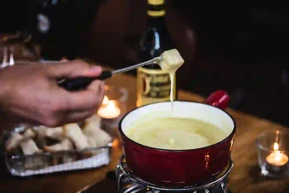 Auf dem Bild ist eine Hand, die einen Fonduegabel mit geschmolzenem Käse über einem rot emaillierten Caquelon hält, in dem sich ein cremiges Käsefondue befindet. Im Hintergrund sind gedämpftes Licht und eine Flasche Kirsch zu erkennen, was auf eine gemütliche und genussvolle Atmosphäre hinweist.