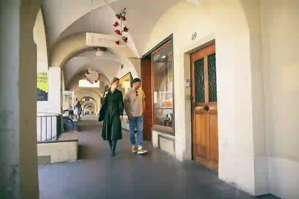 Ein Paar flaniert entspannt durch die Laubengänge der Berner Altstadt, vorbei an lokalen Geschäften und geschmückten Türrahmen, eingehüllt in das historische Flair der Stadt.