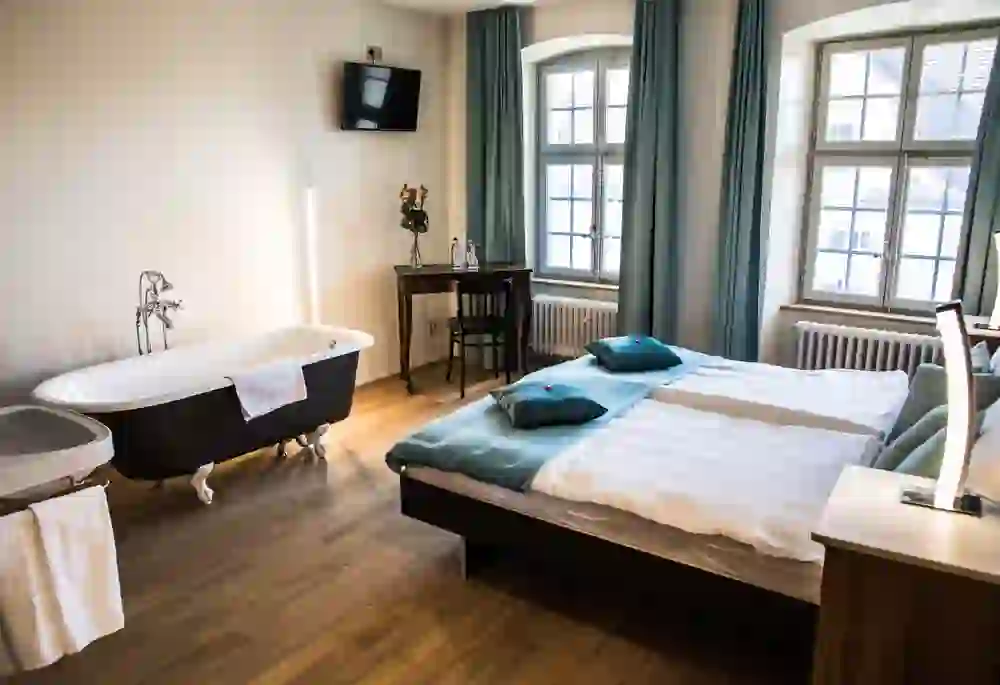 Das Bild zeigt ein helles und einladendes Hotelzimmer mit einer Kombination aus historischem Charme und modernen Elementen. Ein freistehendes Bad in der Ecke, ein großes, gemütliches Bett und klassische Fenster schaffen eine entspannte und luxuriöse Atmosphäre.
