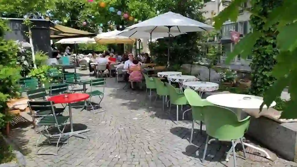L'immagine mostra una terrazza all'aperto del ristorante Ringgenberg Bern. Ci sono diversi tavoli con sedie di diversi colori, distribuiti su un pavimento pavimentato. Grandi ombrelloni fanno ombra. Alcuni ospiti sono seduti ai tavoli, ovviamente godendosi il tempo soleggiato. Sullo sfondo si vedono alberi e un edificio con la bandiera svizzera. Tra gli alberi sono appese delle lanterne decorative.