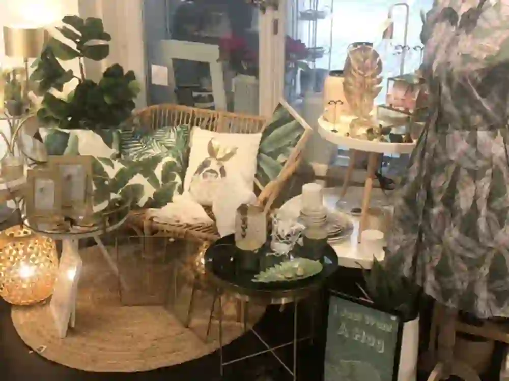 The picture shows a lovingly furnished interior of a decoration or furnishing shop. You can see a rattan armchair with decorative cushions, a round table with various decorative objects and a floor lamp. The arrangement is complemented by plants and candles, creating a cosy and inviting atmosphere.