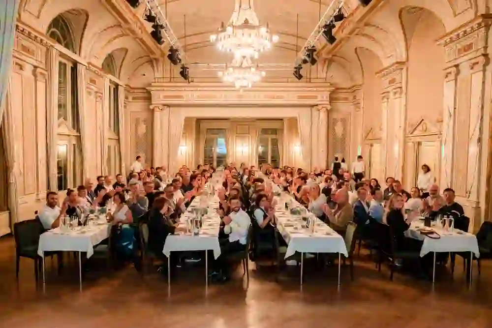 Das Bild zeigt ein elegantes Galadinner in einem prunkvollen, historischen Saal mit hohen Decken und Kronleuchtern. Gäste sitzen an langen, festlich gedeckten Tafeln, applaudieren und wirken gut gelaunt. Die Atmosphäre ist festlich, stilvoll und gemeinschaftlich.