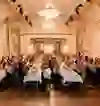 La photo montre un élégant dîner de gala dans une somptueuse salle historique aux hauts plafonds et aux lustres. Les invités sont assis à de longues tables dressées pour la fête, applaudissent et semblent de bonne humeur. L'ambiance est festive, stylée et communautaire.