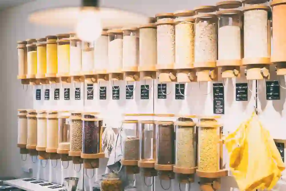 L'image montre un concept de magasin moderne et durable avec un mur de distributeurs pour différents aliments en vrac comme les céréales, les graines et les légumineuses. Les récipients transparents sont disposés de manière ordonnée en rangées et sont munis d'étiquettes indiquant le nom et le prix de leur contenu. Il s'agit d'une partie d'un magasin sans emballage ou d'une épicerie bio où les clients peuvent remplir leurs propres récipients afin de réduire les déchets d'emballage.