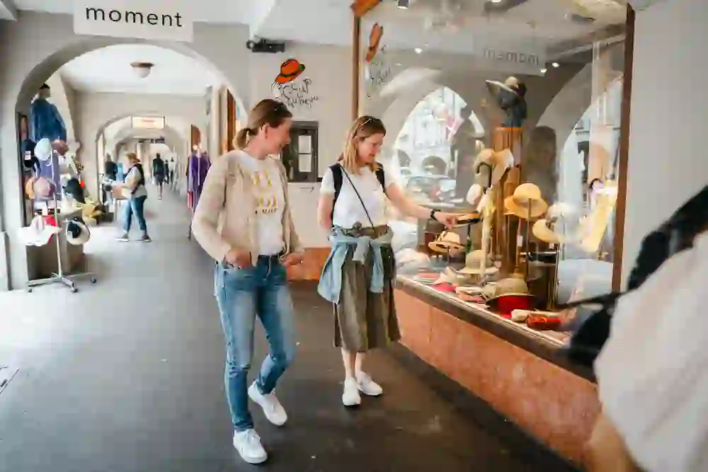 L'image montre deux femmes qui se tiennent devant un magasin de chapeaux dans une arcade très fréquentée et qui regardent à travers la vitrine. L'une d'elles désigne un chapeau et semble intéressée. L'atmosphère est détendue et joyeuse, elles apprécient d'explorer ensemble les magasins et de passer des moments de détente en flânant.
