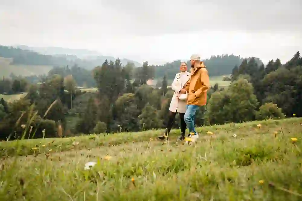 Das Bild zeigt ein Paar, das fröhlich durch eine grüne Hügellandschaft spaziert. Die beiden lachen und genießen den Moment, umgeben von wilder Natur und frischen Wiesenblumen. Die Stimmung ist entspannt und romantisch, voller Freude an der gemeinsamen Zeit im Freien.
