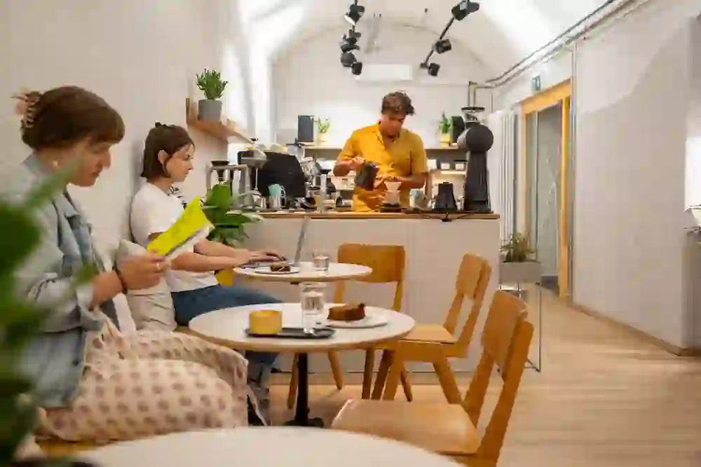 Im Café Friend or Foe in Bern genießen Menschen in entspannter Atmosphäre ihren Kaffee. Eine Person liest, eine andere arbeitet am Laptop, während der Barista im Hintergrund Kaffee zubereitet. Die ruhige, gemütliche Stimmung lädt zum Verweilen und Entspannen ein, ideal für eine kleine Auszeit im Alltag. Die warme Beleuchtung und das minimalistische Design schaffen eine harmonische und inspirierende Umgebung.