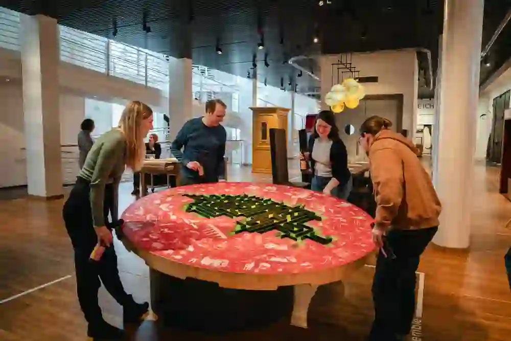 The picture shows an interactive exhibition at the Museum of Communication in Bern. Four people are standing around a large, round table that represents an interactive game or installation. The table has an eye-catching design with red and green elements, and the people appear to be actively involved in the game. They are holding drinks in their hands and appear concentrated and amused. Other exhibits and visitors can be seen in the background. The atmosphere is relaxed and inviting, and the museum seems to be a place where learning and fun are combined.