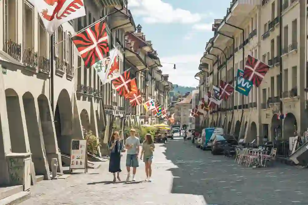 Eine Familie spaziert entspannt durch eine die Berner Altstadt, geschmückt mit bunten Fahnen. Die Sonne taucht die Szenerie in warmes Licht, während Cafés und Geschäfte zum Verweilen einladen. Die Atmosphäre wirkt lebendig, charmant und zugleich entspannt.