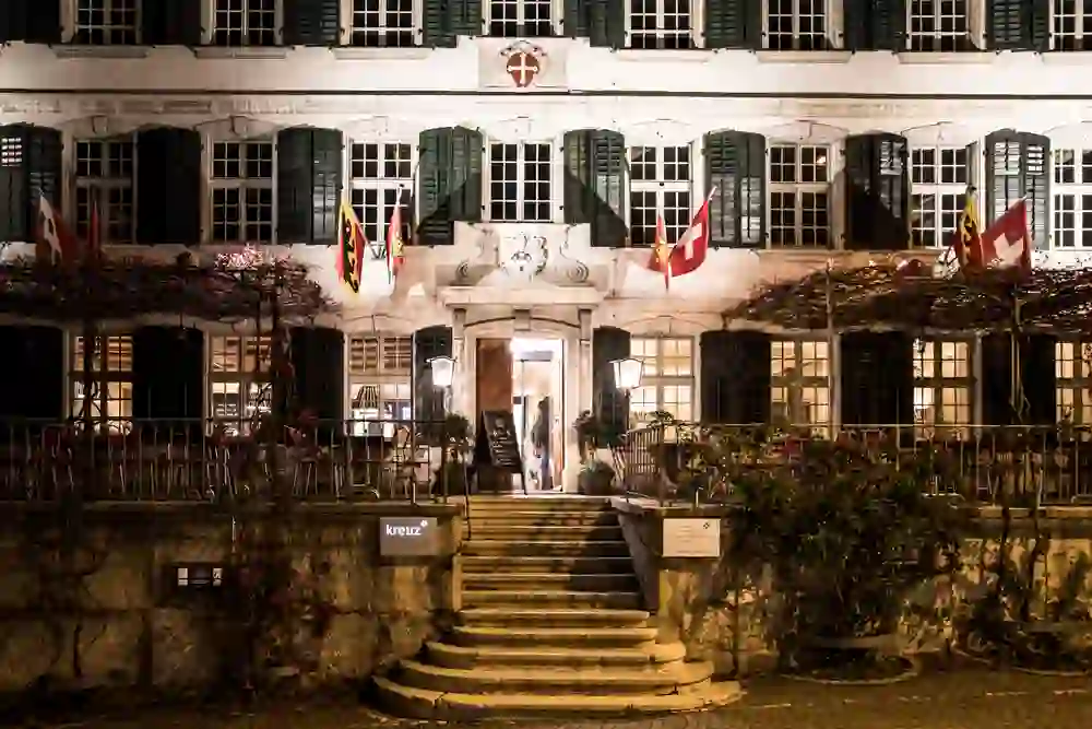 L'image montre la façade nocturne d'un hôtel traditionnel avec des fenêtres éclairées et un escalier menant à une entrée avec des portes ouvertes. Des drapeaux ornent le mur extérieur et l'éclairage confère au lieu une atmosphère chaleureuse et accueillante.