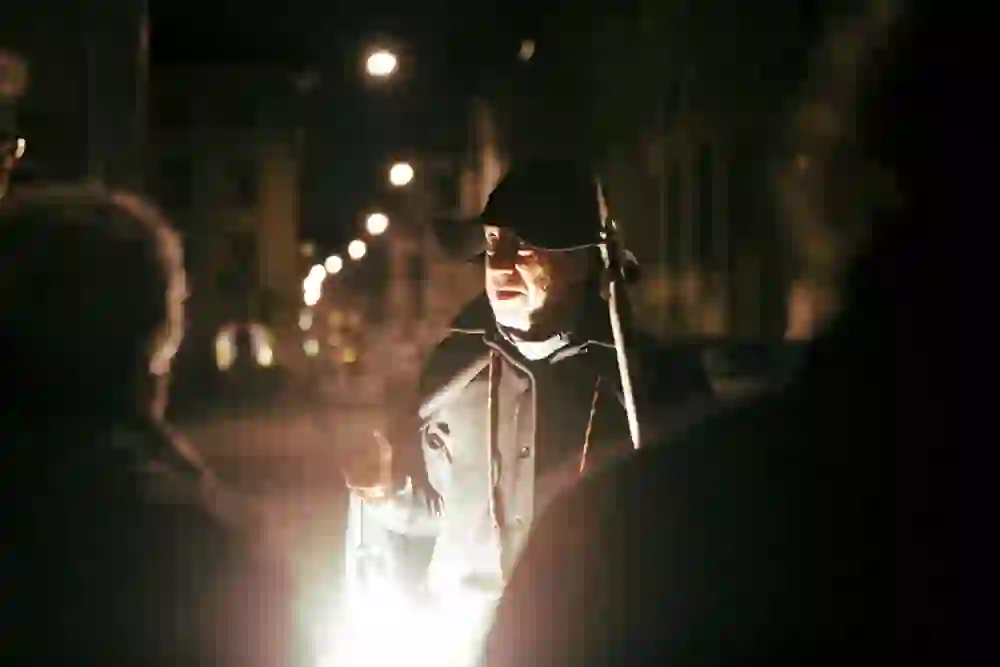 Der Nachtwächter, beleuchtet von seiner Laterne, scheint eine Geschichte zu erzählen, seine Zuhörer sind im Gegenlicht nur als Silhouetten zu erkennen. Sein Gesichtsausdruck ist engagiert, fast theatralisch, was die dramatische Stimmung unterstreicht, die von den warmen Lichtern und dem nächtlichen Hintergrund verstärkt wird.