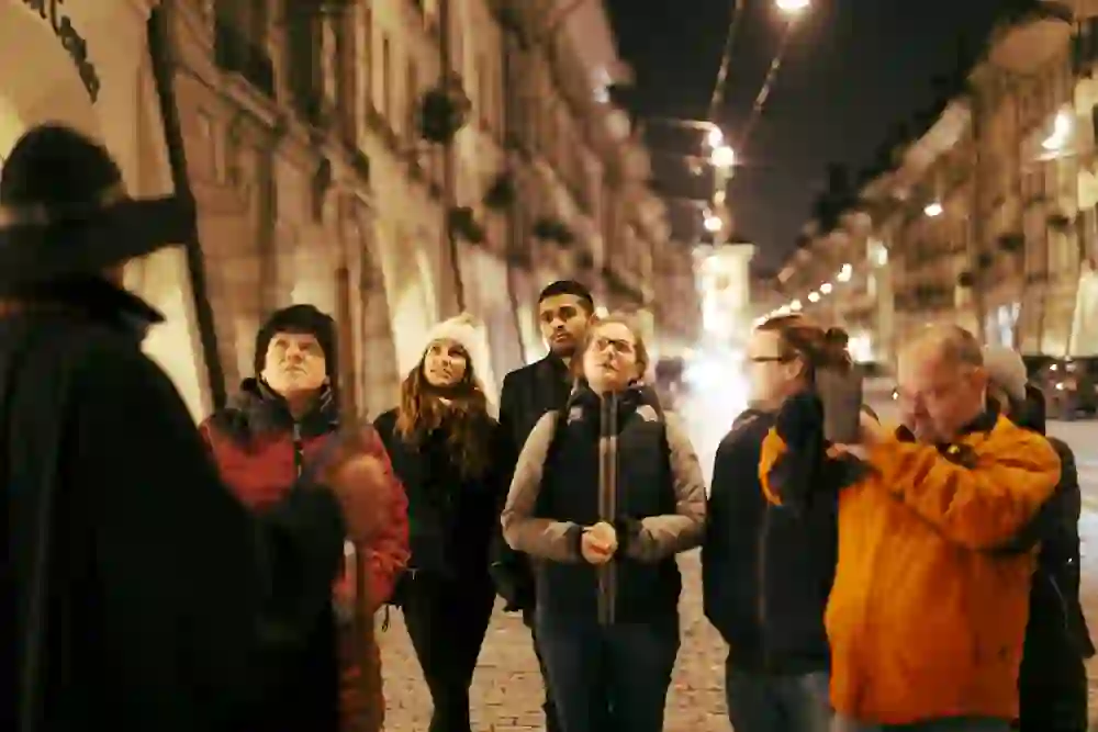 Auf dem Bild ist eine Gruppe von Menschen zu sehen, die aufmerksam einem verkleideten Nachtwächter bei einer abendlichen Stadtführung zuhören. Die Personen wirken fasziniert und eingehüllt in das warme Licht der Strassenlaternen, was eine gemütliche und neugierige Atmosphäre schafft.
