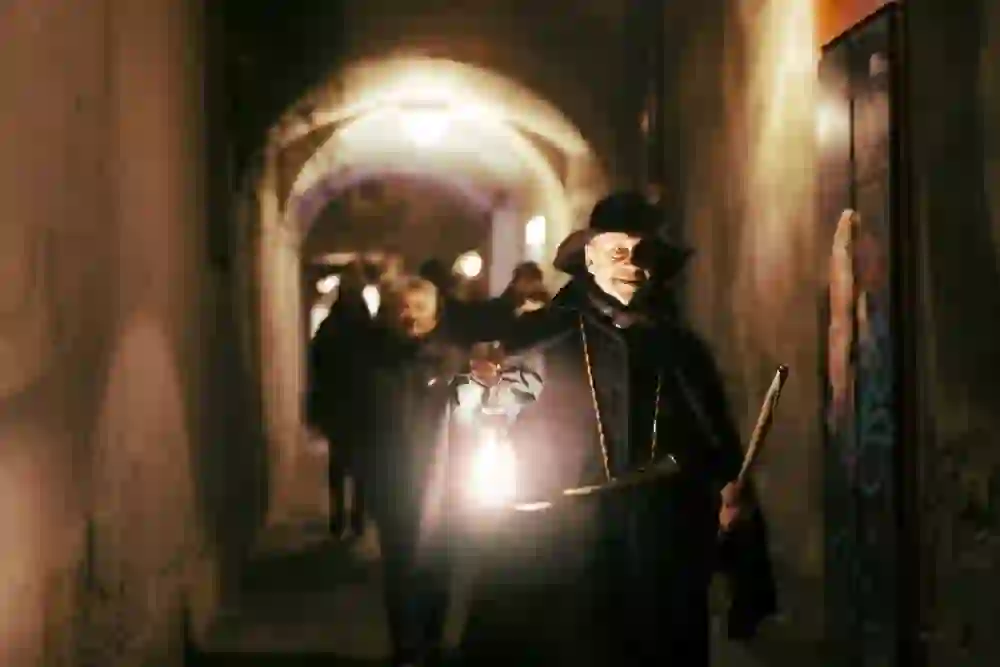 Das Bild zeigt eine nächtliche Szene mit einem Mann, der als Nachtwächter verkleidet ist und eine Laterne hält, während er eine Gruppe durch enge, gewölbte Gänge führt. Die Beleuchtung wirkt warm und geheimnisvoll, und der Nachtwächter blickt direkt in die Kamera, was ihm eine eindringliche Präsenz verleiht. Es ist eine Stimmung von Nostalgie und Spannung spürbar.