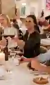 Eine fröhliche Gruppe junger Erwachsener sitzt an einem festlich gedeckten Tisch in einem stilvollen Restaurant. Sie stossen mit Gläsern Rotwein an und geniessen gemeinsam ein mehrgängiges Menü. Die Atmosphäre ist warm und gesellig, im Hintergrund sitzen weitere Gäste zwischen kunstvoll dekorierten Wänden.