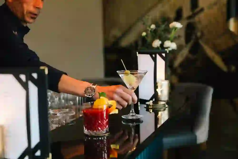 L'immagine mostra un barista in piedi davanti a un elegante bancone che sta per servire un cocktail. L'ambiente ha un'atmosfera accogliente, con luci soffuse e fiori sullo sfondo, che fa pensare a un bar o a una lounge di lusso.