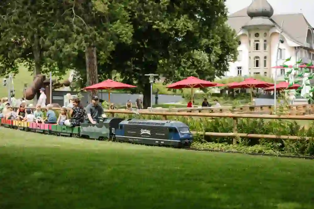 Das Bild zeigt eine fröhliche Szene im Gurtenpark: Kinder und Erwachsene fahren mit einem Miniaturzug durch eine grüne, sonnendurchflutete Parklandschaft, vorbei an entspannten Cafébesuchern unter roten Sonnenschirmen, die das gesellige Beisammensein geniessen.