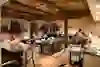 The picture shows a lively restaurant with people sitting and eating at different tables. The lighting from stylish hanging lamps and the visible wooden beam interior create a warm and cosy atmosphere. The blurred movement of the guests conveys a dynamic mood.