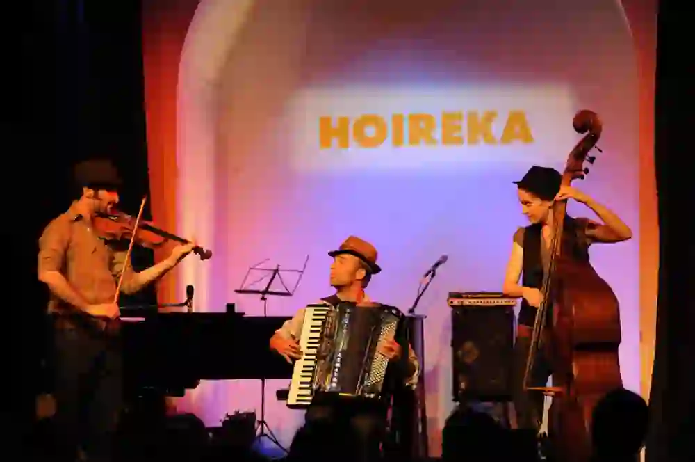Das Bild zeigt drei Musiker, die leidenschaftlich auf ihren Instrumenten spielen – Violine, Akkordeon und Kontrabass – vor einem Hintergrund mit dem Schriftzug "HOIREKA". Ihre Hüte und das warme Bühnenlicht kreieren eine stimmungsvolle und gemütliche Atmosphäre, typisch für einen Abend im La Cappella.