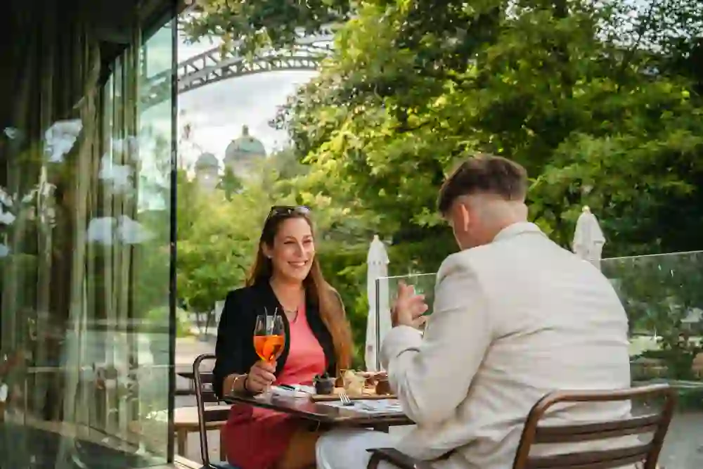 Zwei Menschen sitzen auf einer sonnigen Terrasse und geniessen Getränke und Snacks. Die Frau lacht, während der Mann gestikuliert. Im Hintergrund ist das Bundeshaus, Bäume und eine Brücke zu sehen. Die Stimmung ist entspannt, freundlich und voller Sommerfreude.
