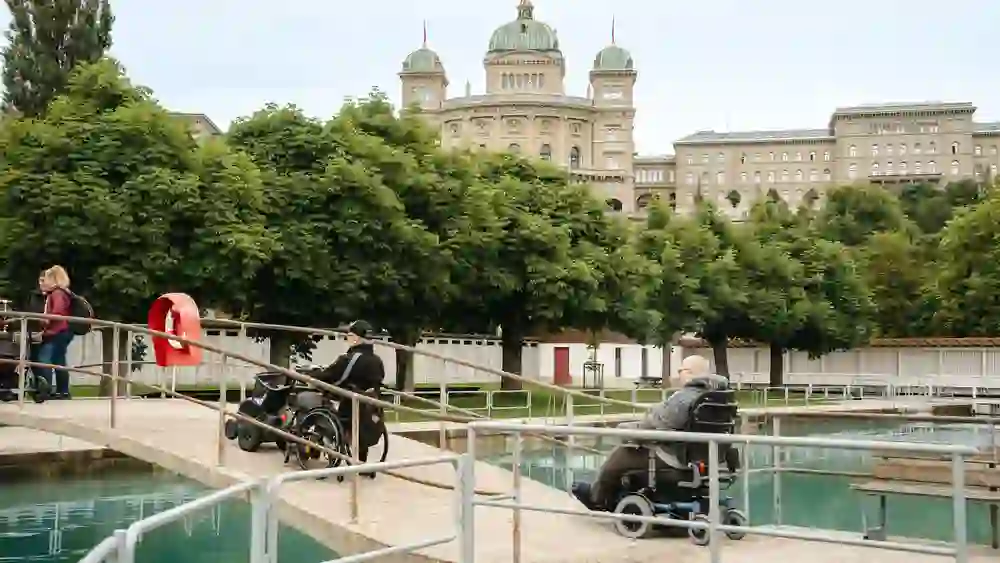 Das Bild zeigt eine Gruppe von Menschen, die einen sonnigen Tag im Freibad Marzili genießen, mit dem Bundeshaus im Hintergrund. Rollstuhlfahrer und Fußgänger teilen sich friedlich die Brücke, was eine inklusive und entspannte Atmosphäre vermittelt.