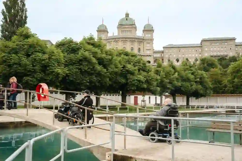 Das Bild zeigt eine Gruppe von Menschen, die einen sonnigen Tag im Freibad Marzili genießen, mit dem Bundeshaus im Hintergrund. Rollstuhlfahrer und Fußgänger teilen sich friedlich die Brücke, was eine inklusive und entspannte Atmosphäre vermittelt.