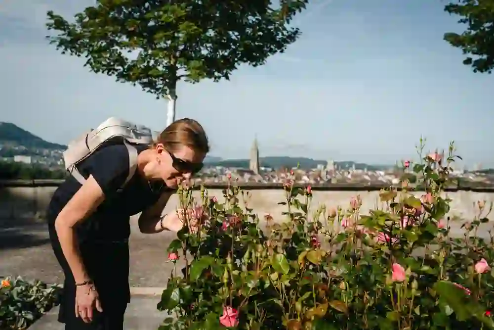 Das Bild zeigt eine Frau, die an Rosen schnuppert, mit Berns Skyline im Hintergrund. Die Stimmung ist ruhig und entspannt, ein Moment der Verbundenheit mit der Natur. Die warme Sonne und die blühenden Rosen schaffen eine Atmosphäre von Freude und Gelassenheit.